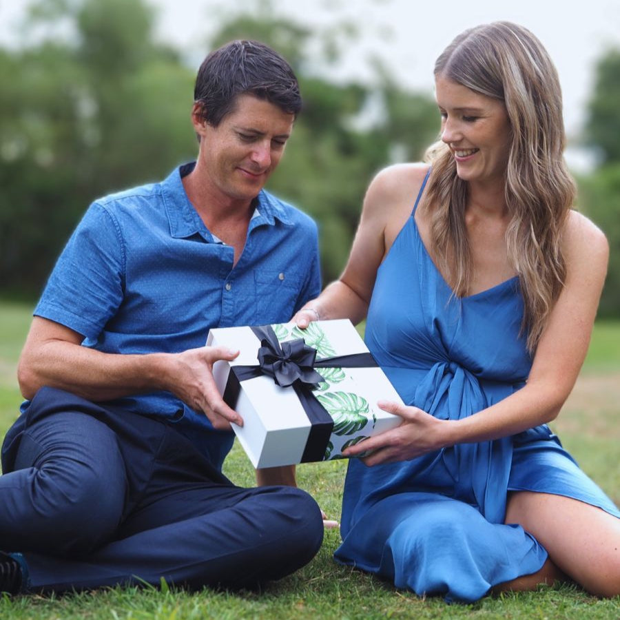 Smiling couple exchanging a beautifully wrapped wickedly indulgent gift hamper in a lush Invercargill park — perfect for birthdays, thank-yous, and just-because moments with free NZ delivery over $150.