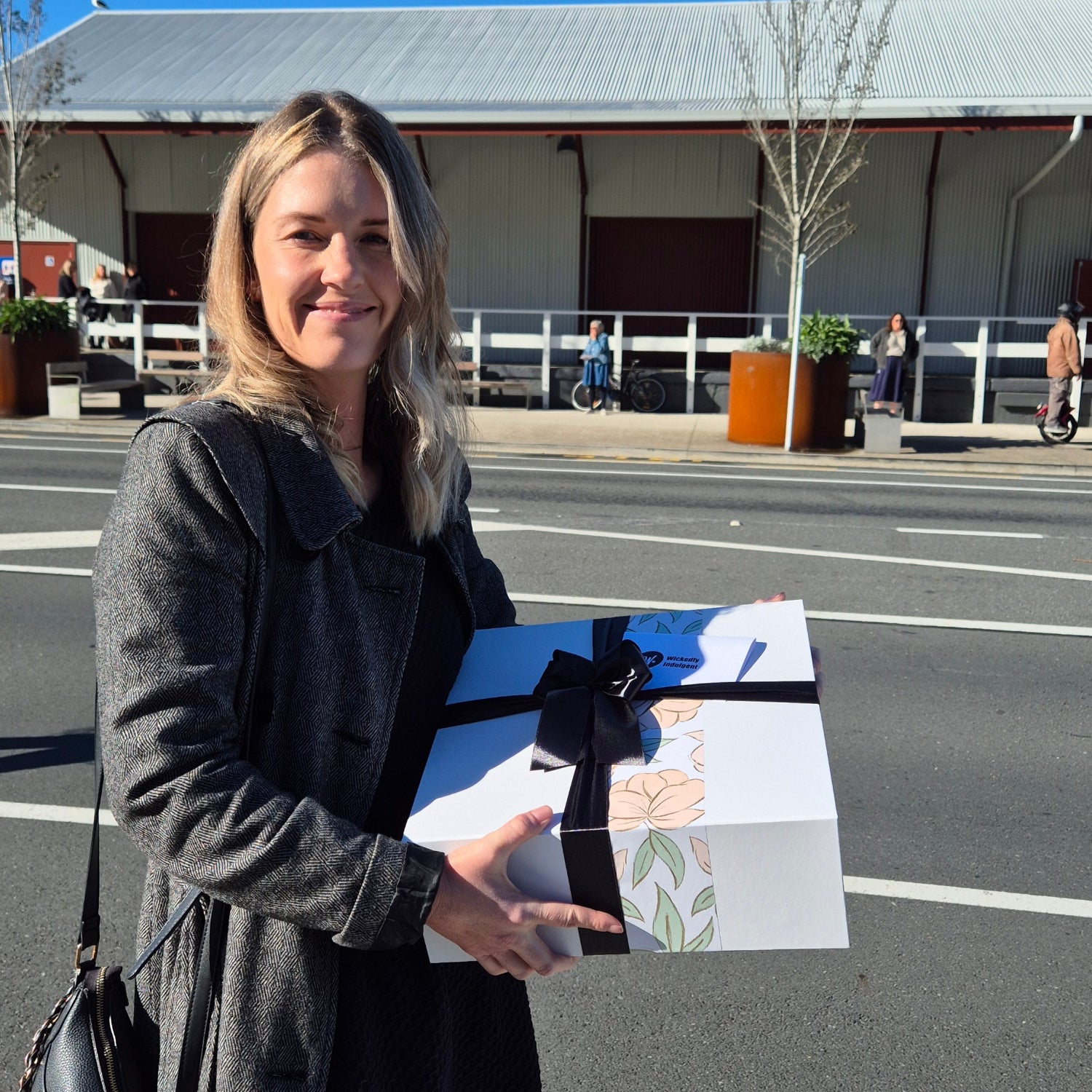 Smiling woman holding a ribbon-wrapped Wickedly Indulgent gift hamper on a sunny Auckland street