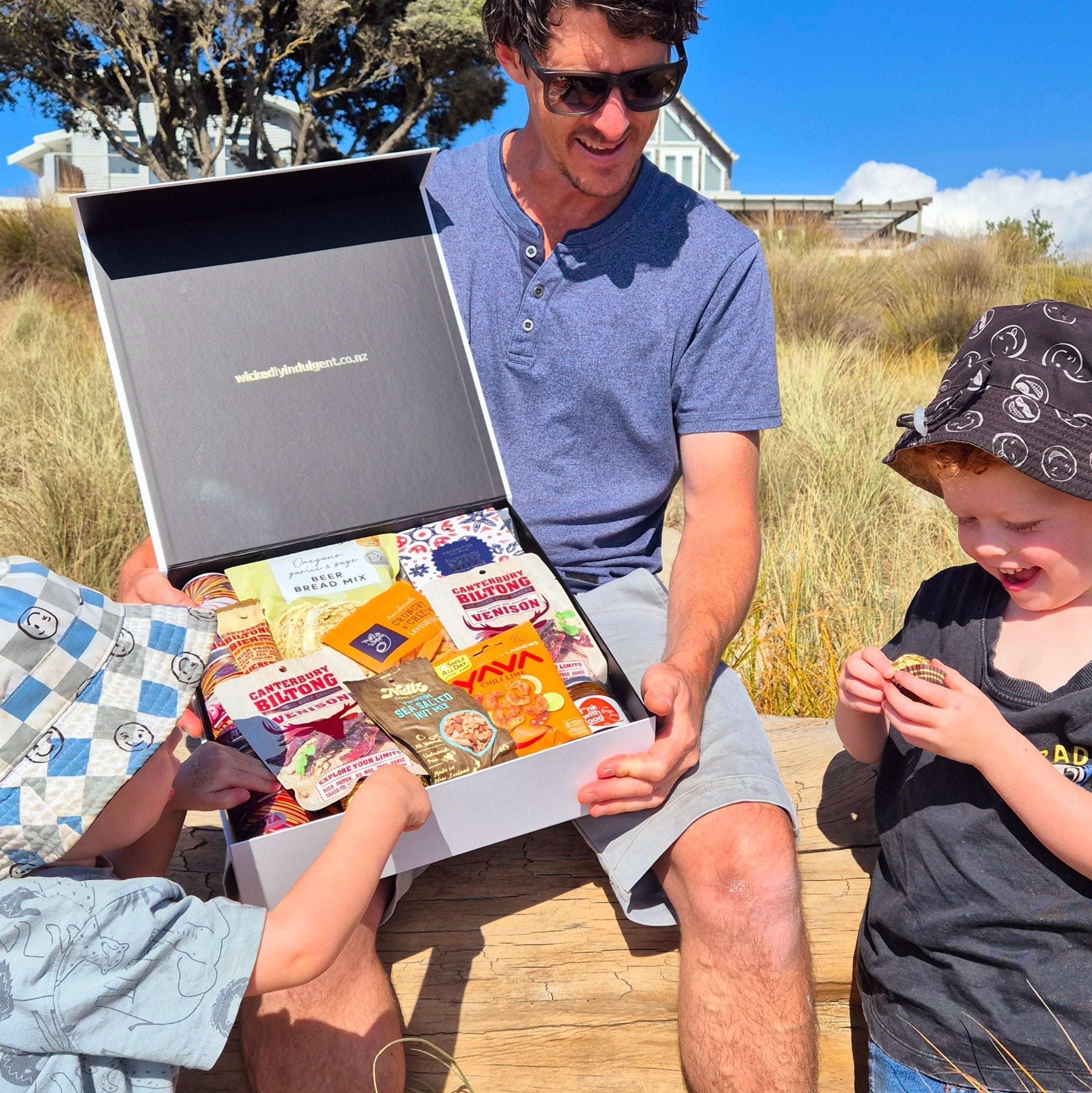 Father opening a Wickedly Indulgent gift hamper outdoors with two smiling children, sharing snacks together in the sun on Father’s Day.