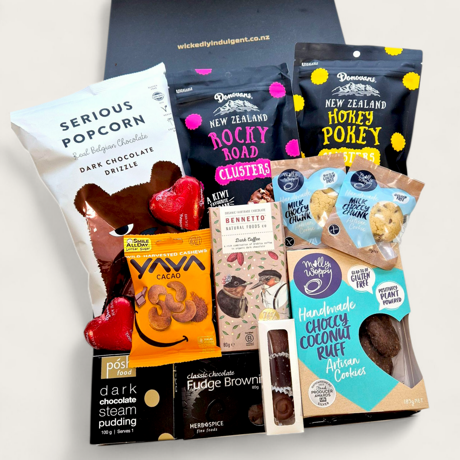 A premium gift hamper called "For Choc’s Sake" featuring chocolate treats including popcorn, cookies, rocky road, artisan bars, pudding, and cacao cashews in a sleek black & white Wickedly Indulgent box.