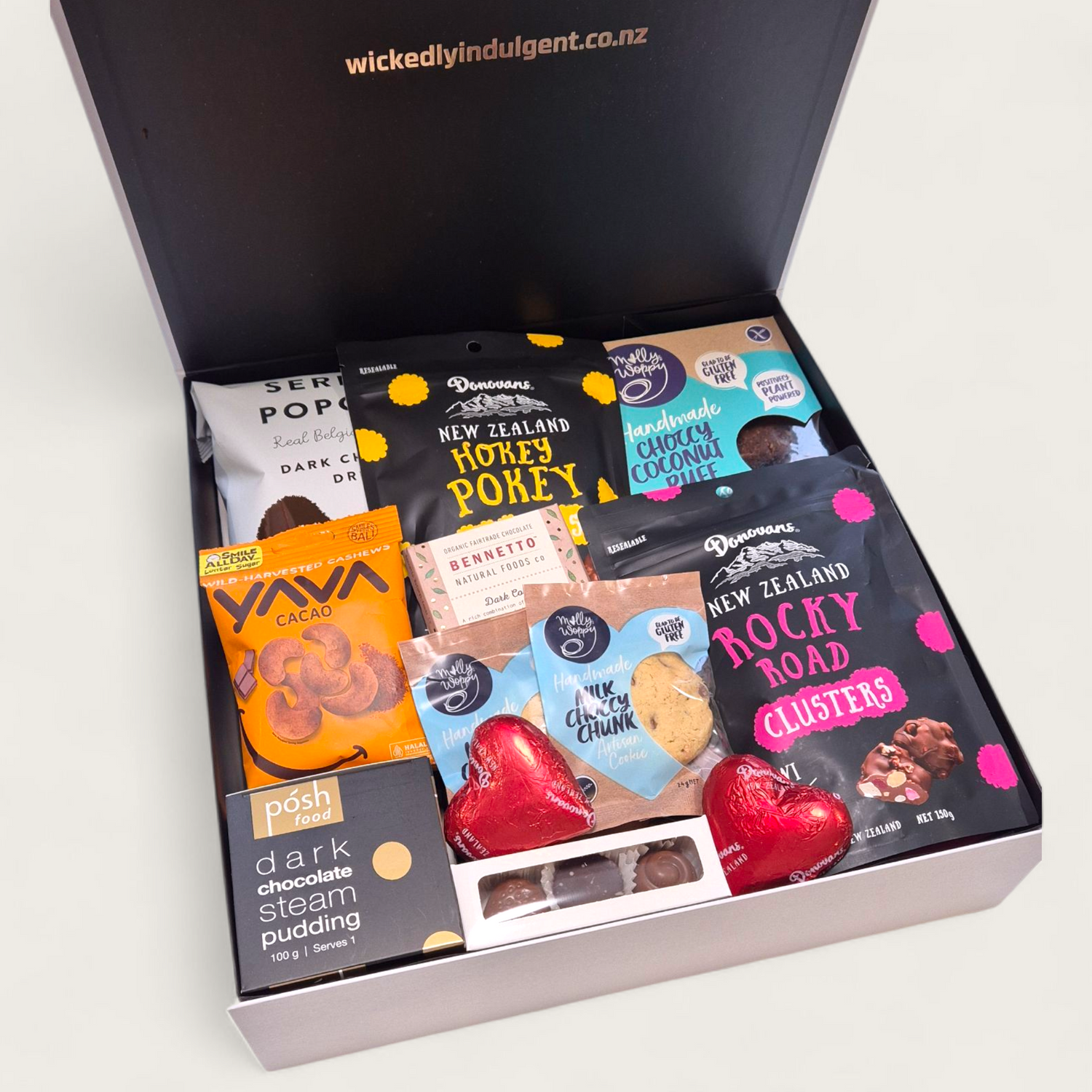 A premium gift hamper called "For Choc’s Sake" featuring chocolate treats including popcorn, cookies, rocky road, artisan bars, pudding, and cacao cashews in a sleek black & white Wickedly Indulgent box.
