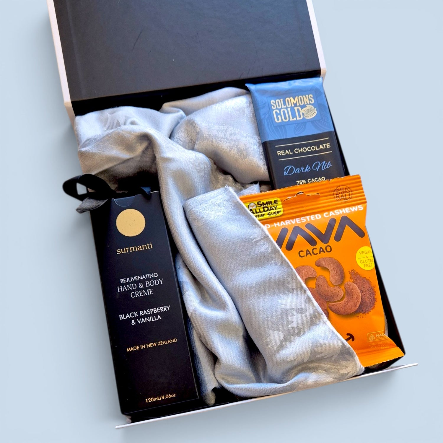 Dairy and gluten free gift box for her containing a blue pashmere scarf, Surmanti natural hand cream, Solomon's Gold vegan chocolate 
tablet, and Yava cacao-coated cashews, presented in a sleek black gift  box with satin ribbon, by Wickedly Indulgent, New Zealand
