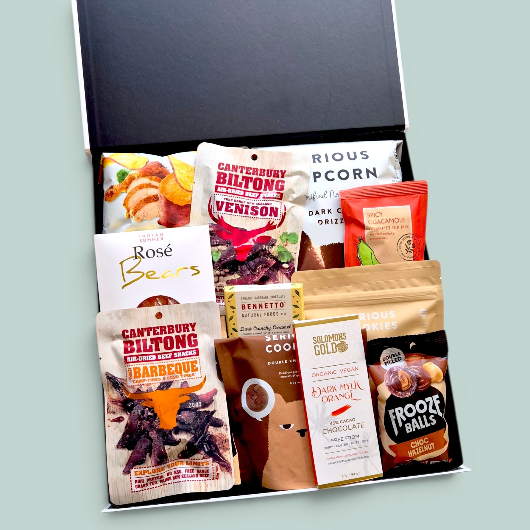 Open black gift box from Wickedly Indulgent NZ filled with Canterbury 
Biltong, Solomon's Gold vegan chocolate, Bennetto chocolate, Indian 
Summer Rosé Bears, Serious Popcorn, Serious Cookies and Frooze Balls 
— GD Treats on Repeat gluten-free snack hamper